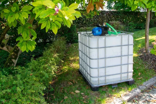 Citerne eau de pluie blanc nettoyé de 1000 litres avec cage galvanisée, pompe, vanne et pallete en plastique ou metal. vue-du-côté-gauche-dans-environnement-de-jardin