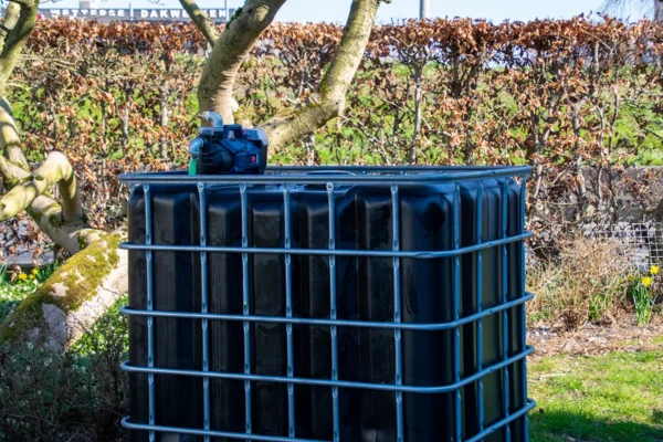 cuve-ibc-noir-avec-pompe-sur-batterie-1000L-renouvele-palette-acier-plastique-vue-de-cote-arrieere-sans-tuyau-sortant-dans-environnement-de-jardin.webp